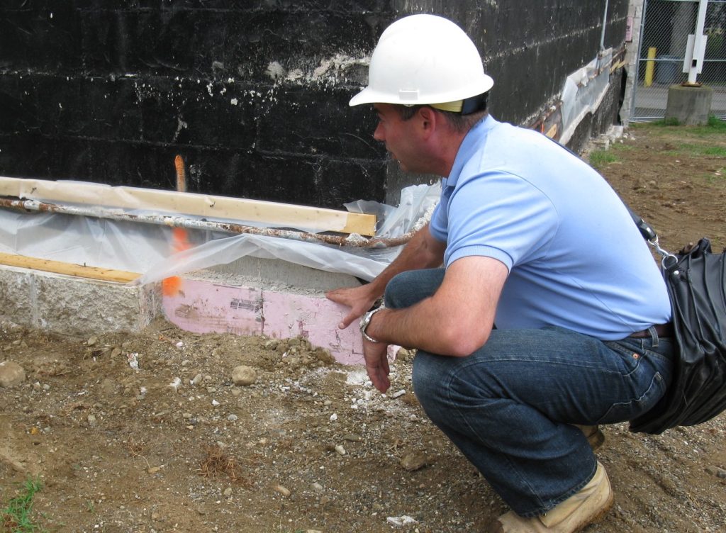 Larry Keenan PE conducts a site visit and squats beside a foundation