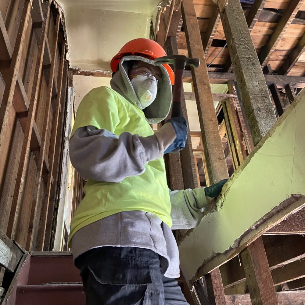 Chelsea Doyle wearing a grey hoodie with a safety green t-shirt on top, an orange hard hat and a protective respirator mask, holding a hammer inside an unfinished building