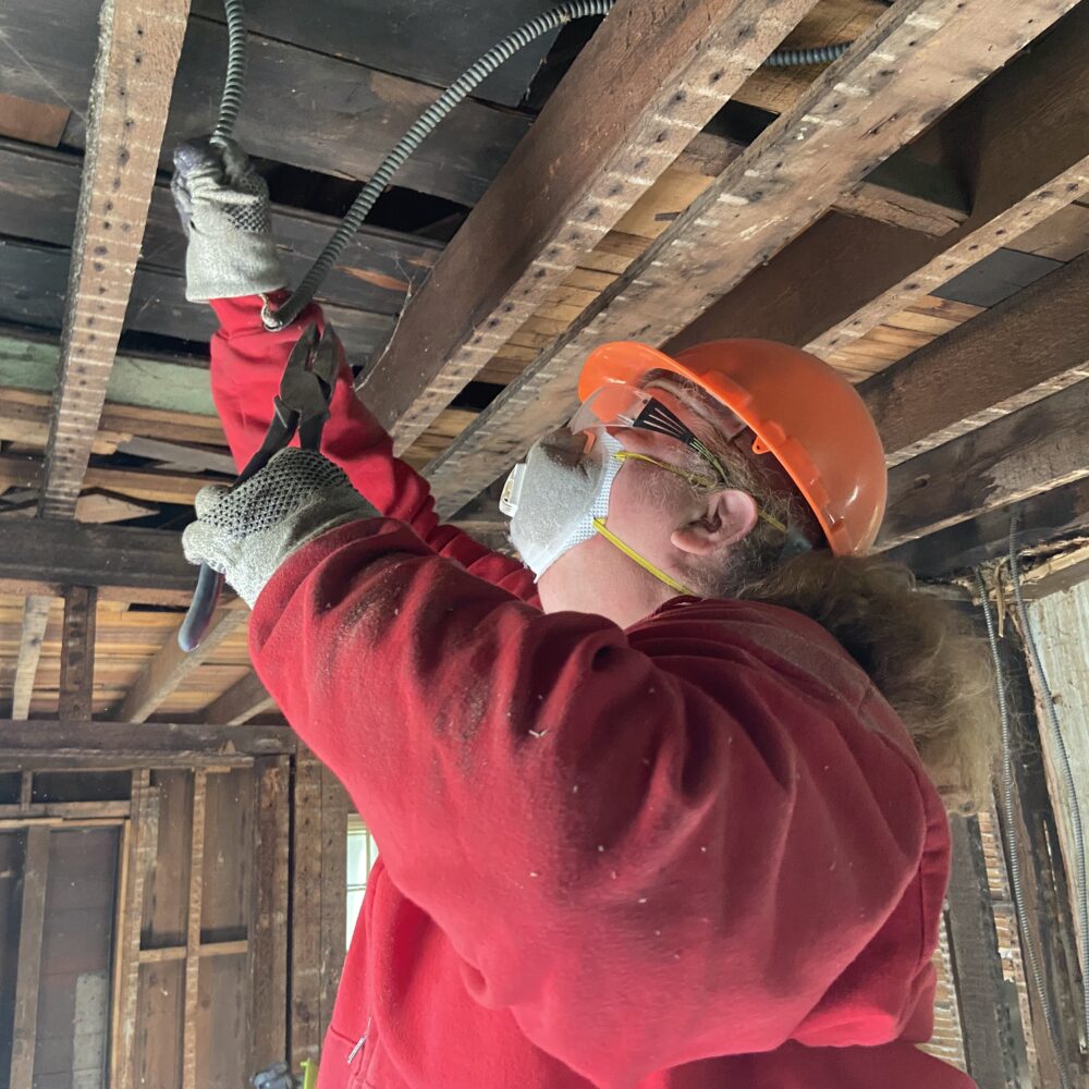 A woman in a respirator mask and hard hat pulling wiring from a ceiling in an unfinished house