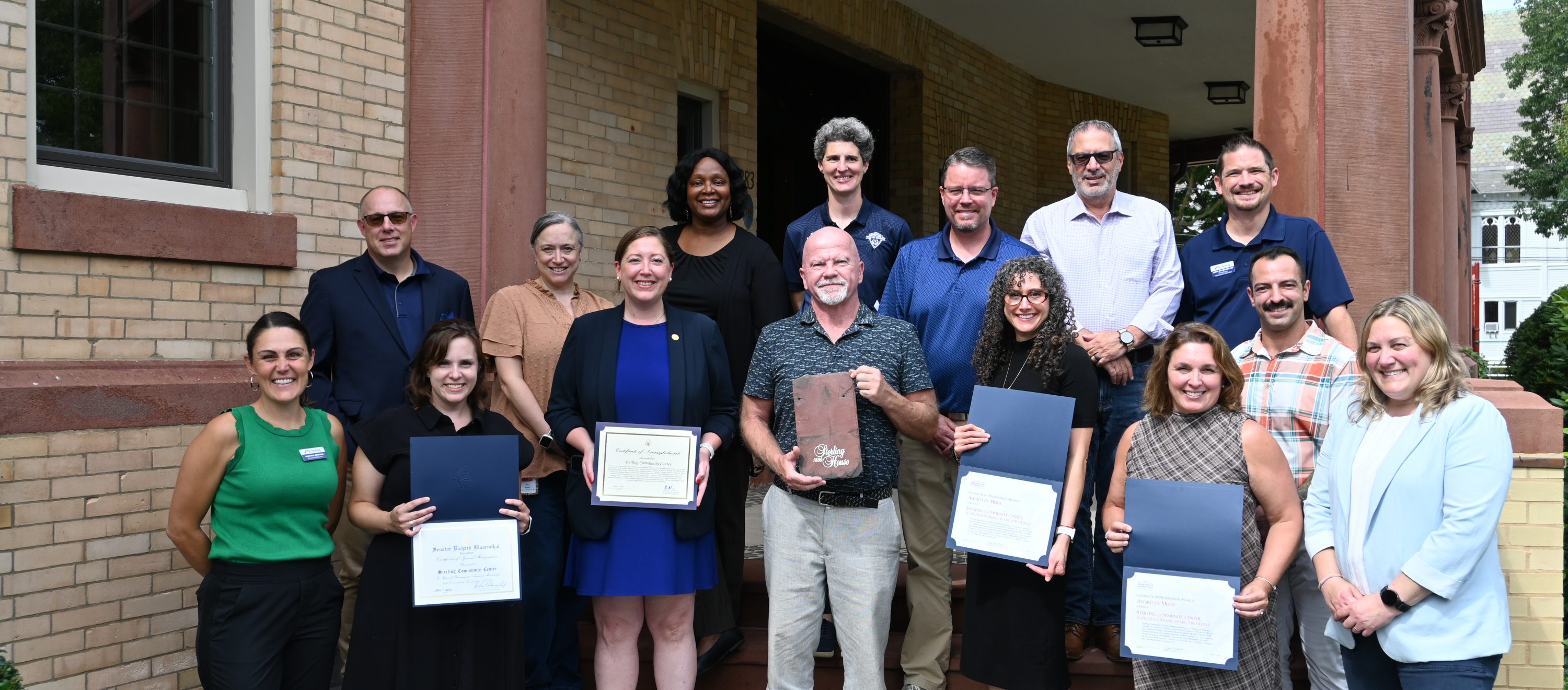 A group posing on exterior stairs holding certificates and plaques