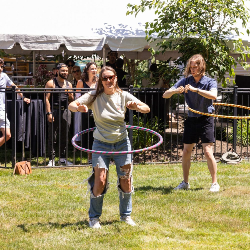 Chelsea Doyle hula-hooping outdoors in front of a white tent with onlookers at a picnic