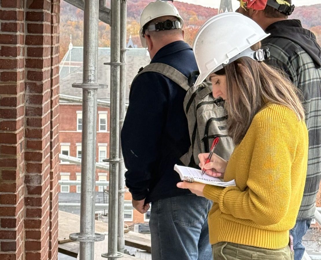 A woman in a hard hat writes in a notebook while standing on a scaffold outside a brick masonry building